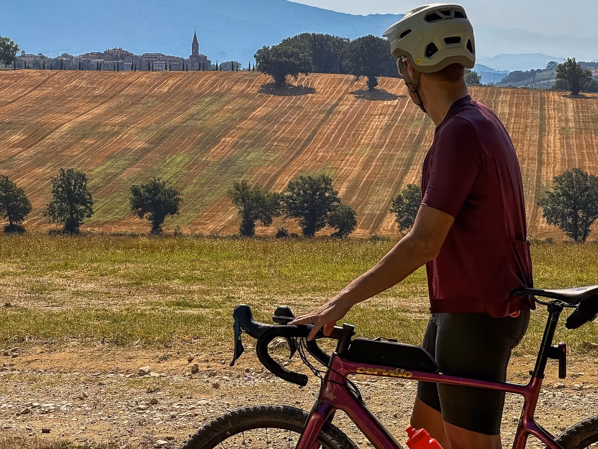 A cyclist standing next to his bike looking over Umbrian fields and hills, with a hilltop village and its bell tower on the horizon